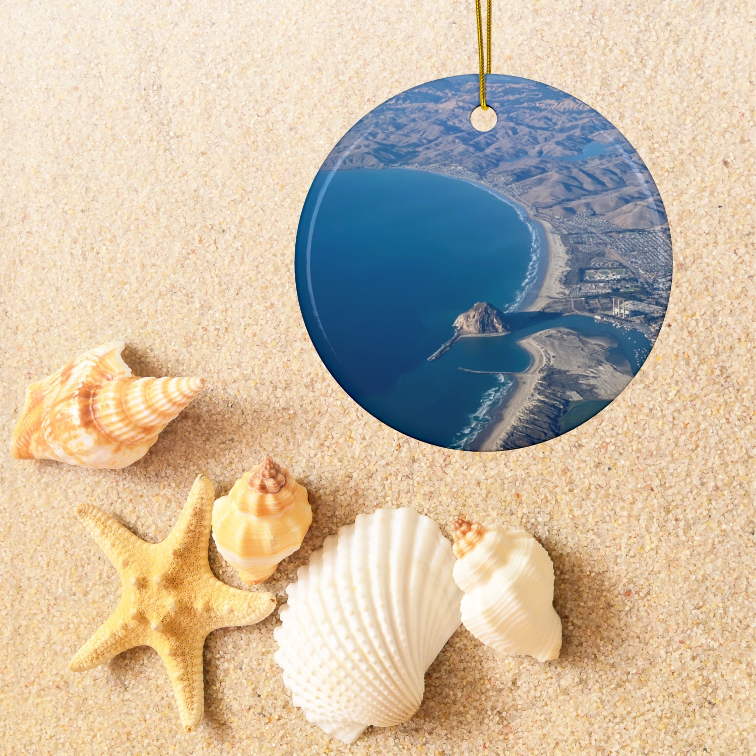 Ceramic holiday ornament featuring an aerial photograph of Morro Rock and the coastline of Morro Bay, California, with vibrant blue water and golden hills, surrounded by seashells and a starfish on sand.