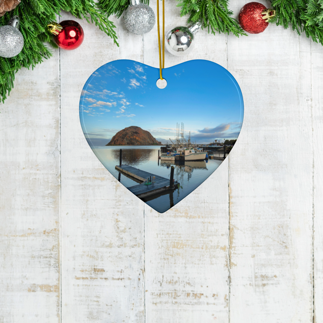 Heart-shaped ceramic Christmas ornament featuring an early morning photograph of Morro Bay harbor with boats, a pier, and Morro Rock reflected in the calm water, hanging on a wooden surface with Christmas decorations.