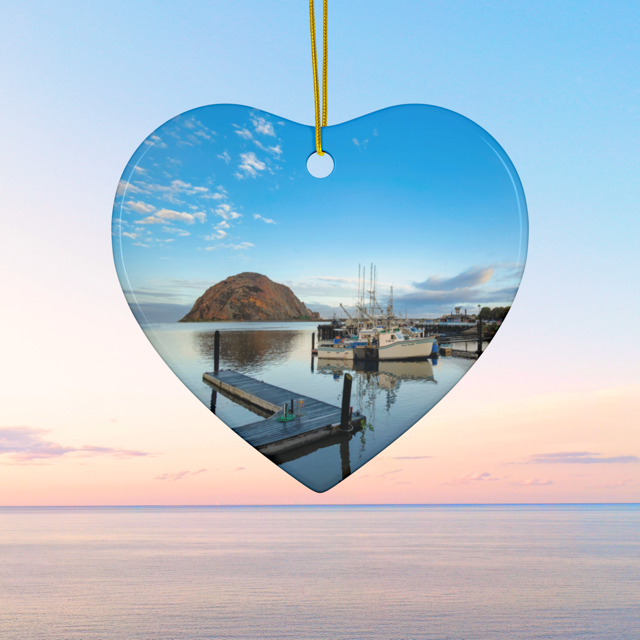Heart-shaped ceramic Christmas ornament featuring an early morning photograph of Morro Bay harbor with boats, a pier, and Morro Rock reflected in the calm water, on a blue sky background