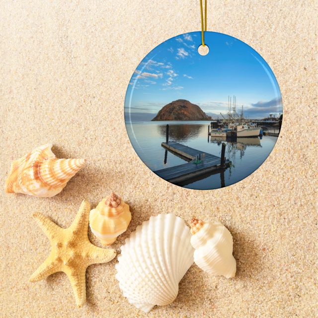 Ceramic Christmas ornament featuring an early morning photograph of Morro Bay harbor with boats, a pier, and Morro Rock reflected in the calm water, surrounded by seashells on sand.