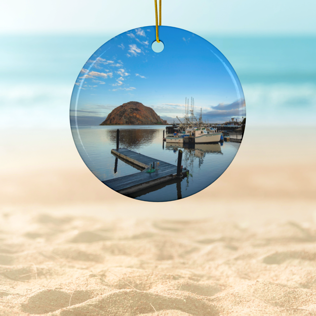 Ceramic Christmas ornament featuring an early morning photograph of Morro Bay harbor with boats, a pier, and Morro Rock reflected in the calm water, on a beach.