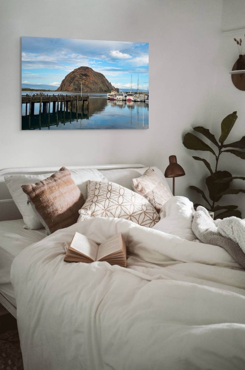 Bedroom with a large canvas print featuring a view of Morro Rock from the harbor in Morro Bay, California with boats and a pier, pillows, and a book.