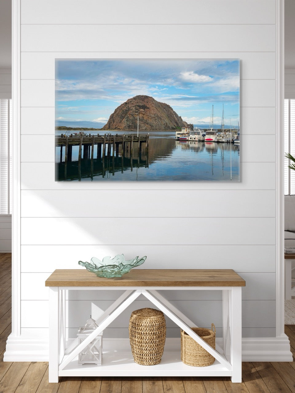 A canvas print featuring a view of Morro Rock from the harbor in Morro Bay, California with boats and a pier in a room with a wooden bench and decorative items.