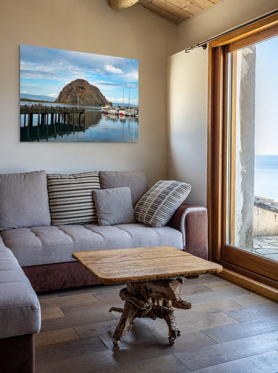 Living room with a gray sofa, wooden coffee table, and a large canvas print featuring a view of Morro Rock from the harbor in Morro Bay, California with boats and a pier.