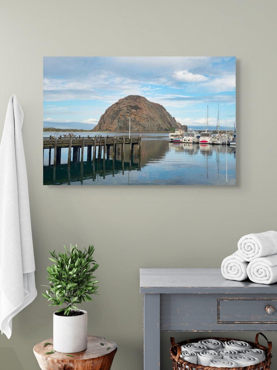 A canvas print featuring a view of Morro Rock from the harbor in Morro Bay, California with boats and a pier on a wall above a dresser.