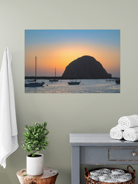 Canvas print of a Morro Rock sunset with sailboats, hanging on a wall above a table with a plant and folded towels.