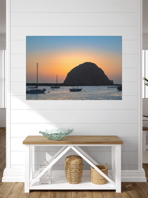 Canvas print of a Morro Rock sunset with boats, displayed on a wall above a wooden bench.