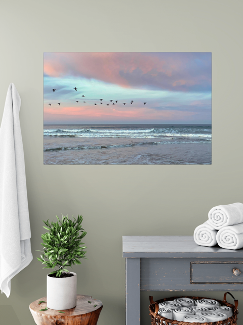 Canvas print of birds flying over pastel pink sunrise in Morro Bay on a wall above a table with a plant and folded towels.