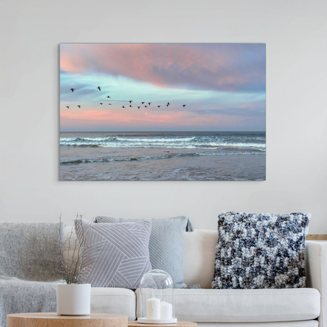 Canvas print of birds flying over pastel pink sunrise in Morro Bay, hanging on a wall above a sofa.