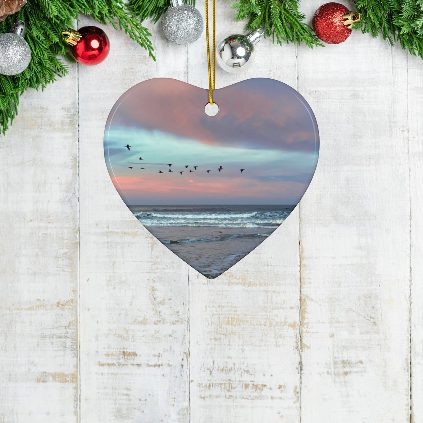 Heart-shaped ceramic holiday ornament featuring a photograph of a flock of birds flying across a pastel pink and violet sunrise sky over the Pacific ocean. on a wooden surface with Christmas decorations.