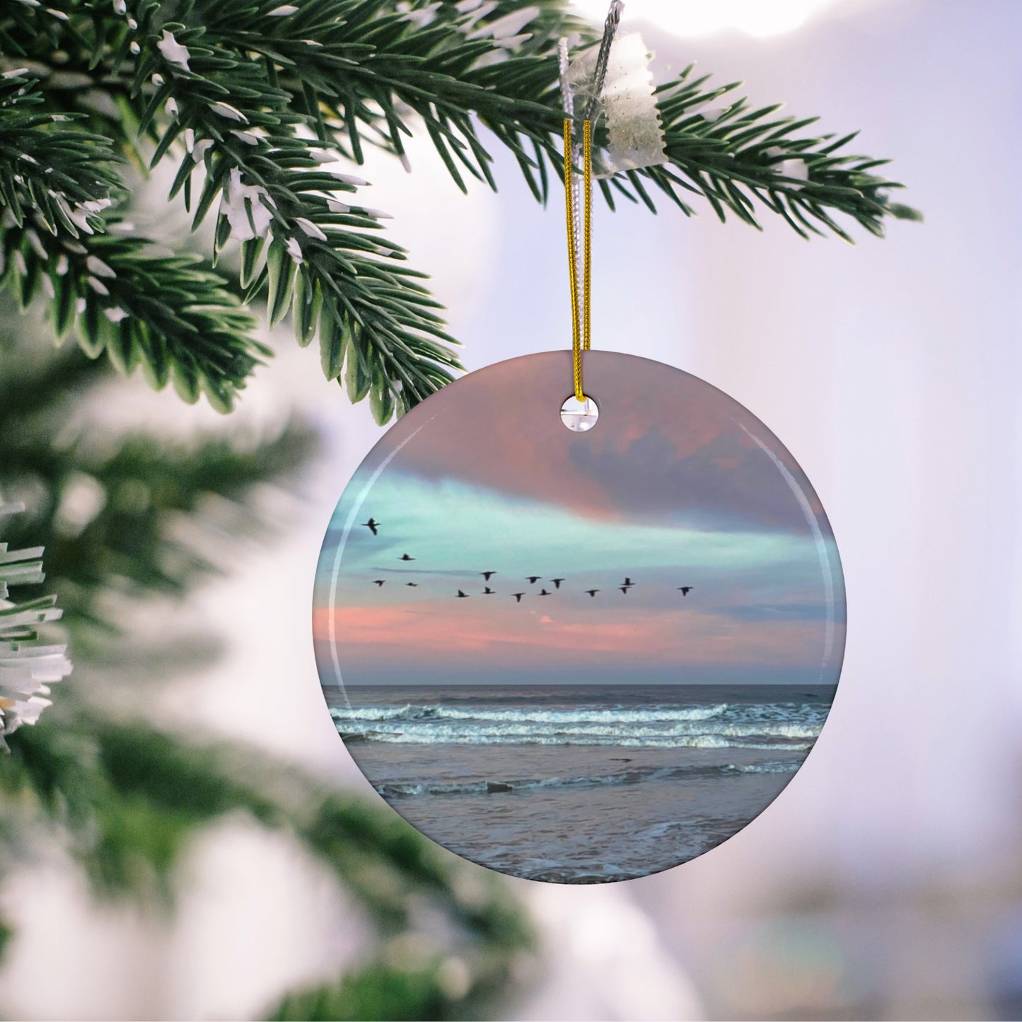 Ceramic holiday ornament featuring a photograph of a flock of birds flying across a pastel pink and violet sunrise sky over the Pacific ocean hanging on a Christmas tree branch