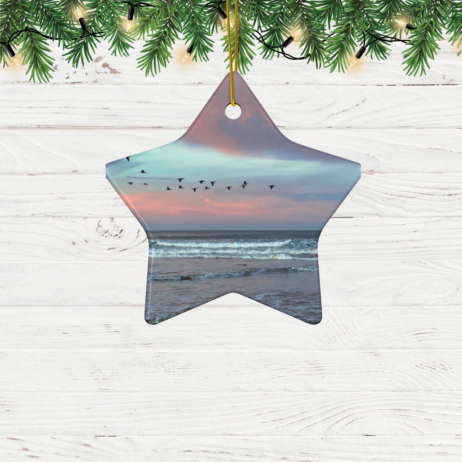Star-shaped oCeramic holiday ornament featuring a photograph of a flock of birds flying across a pastel pink and violet sunrise sky over the Pacific ocean hanging on a wooden surface with Christmas tree branches.
