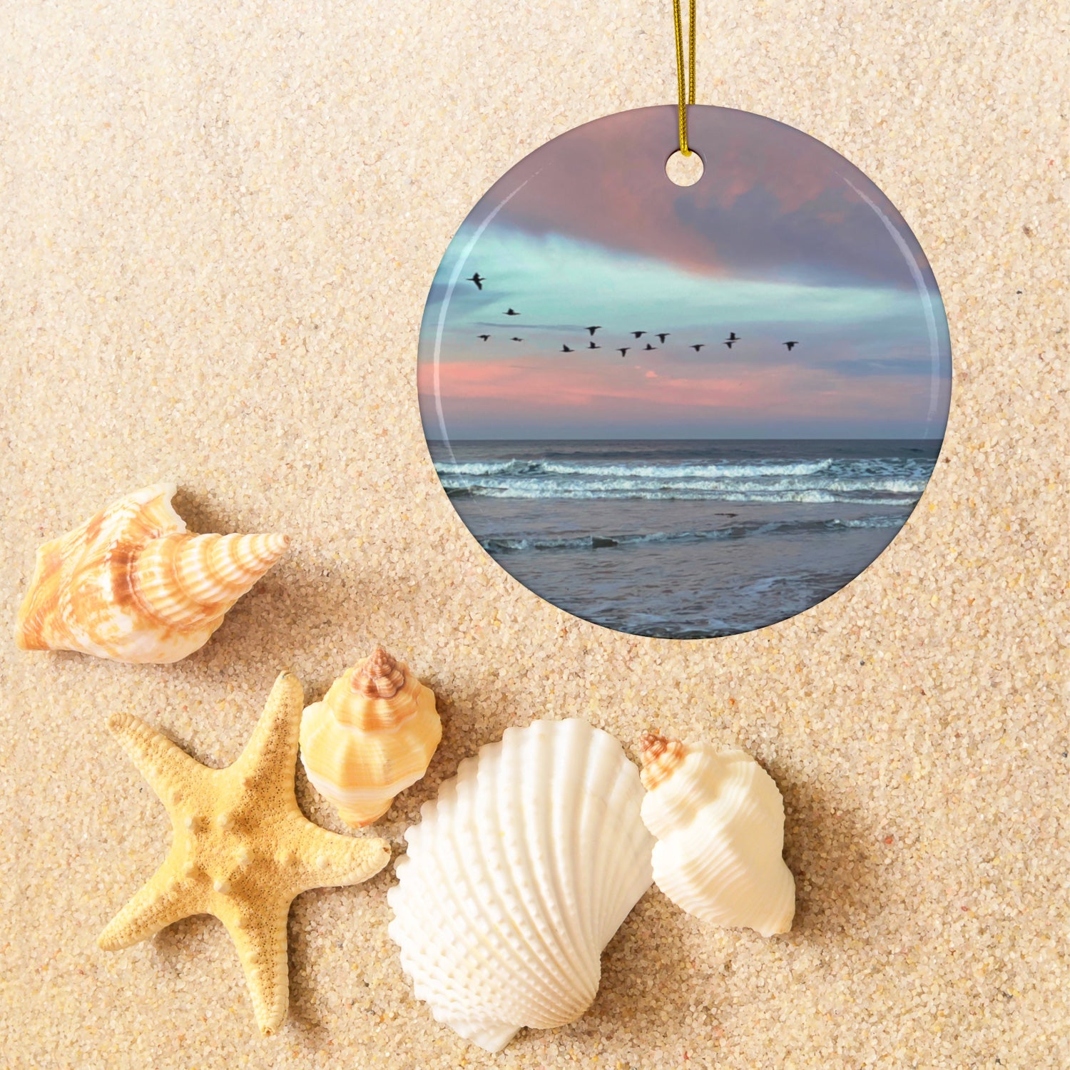 Ceramic holiday ornament featuring a photograph of a flock of birds flying across a pastel pink and violet sunrise sky over the Pacific ocean on sand with shells