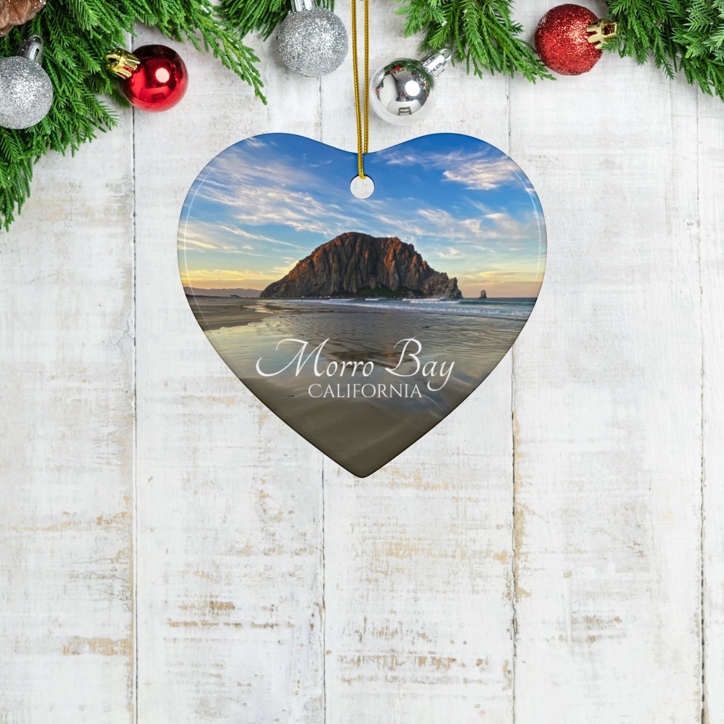 Heart-shaped ceramic holiday ornament featuring a photograph of Morro Rock in Morro Bay, California, with reflections in the sand under a dramatic afternoon sky on a wooden surface with Christmas decorations.