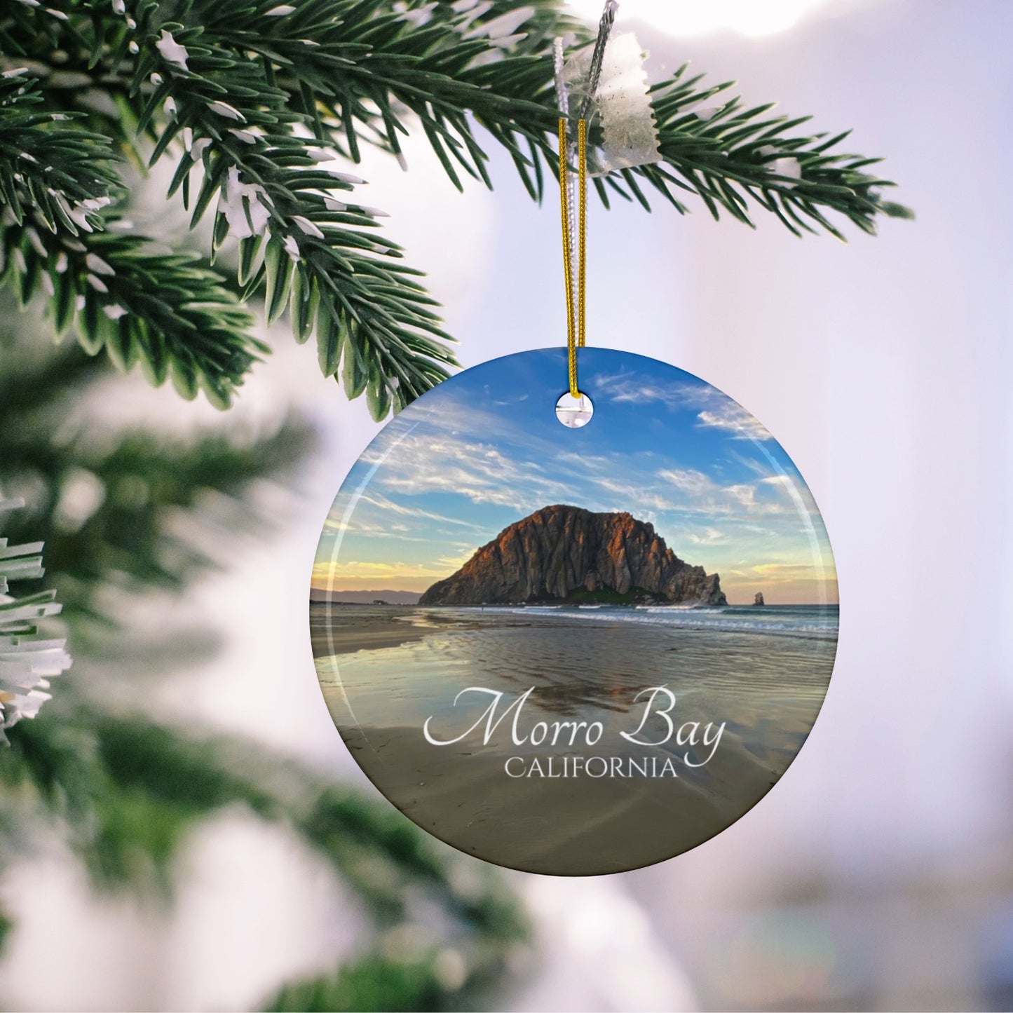 Ceramic holiday ornament featuring a photograph of Morro Rock in Morro Bay, California, with reflections in the sand under a dramatic afternoon sky hanging on a Christmas tree branch.
