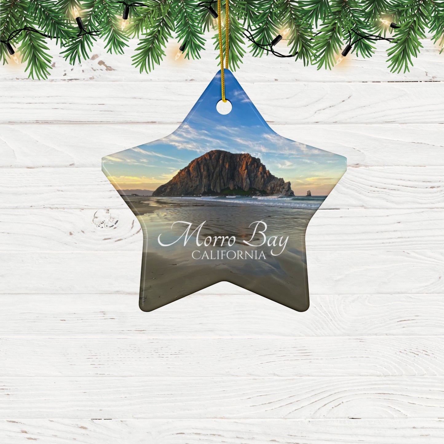 Star-shaped ceramic holiday ornament featuring a photograph of Morro Rock in Morro Bay, California, with reflections in the sand under a dramatic afternoon sky. on a wooden surface with Christmas tree branches.