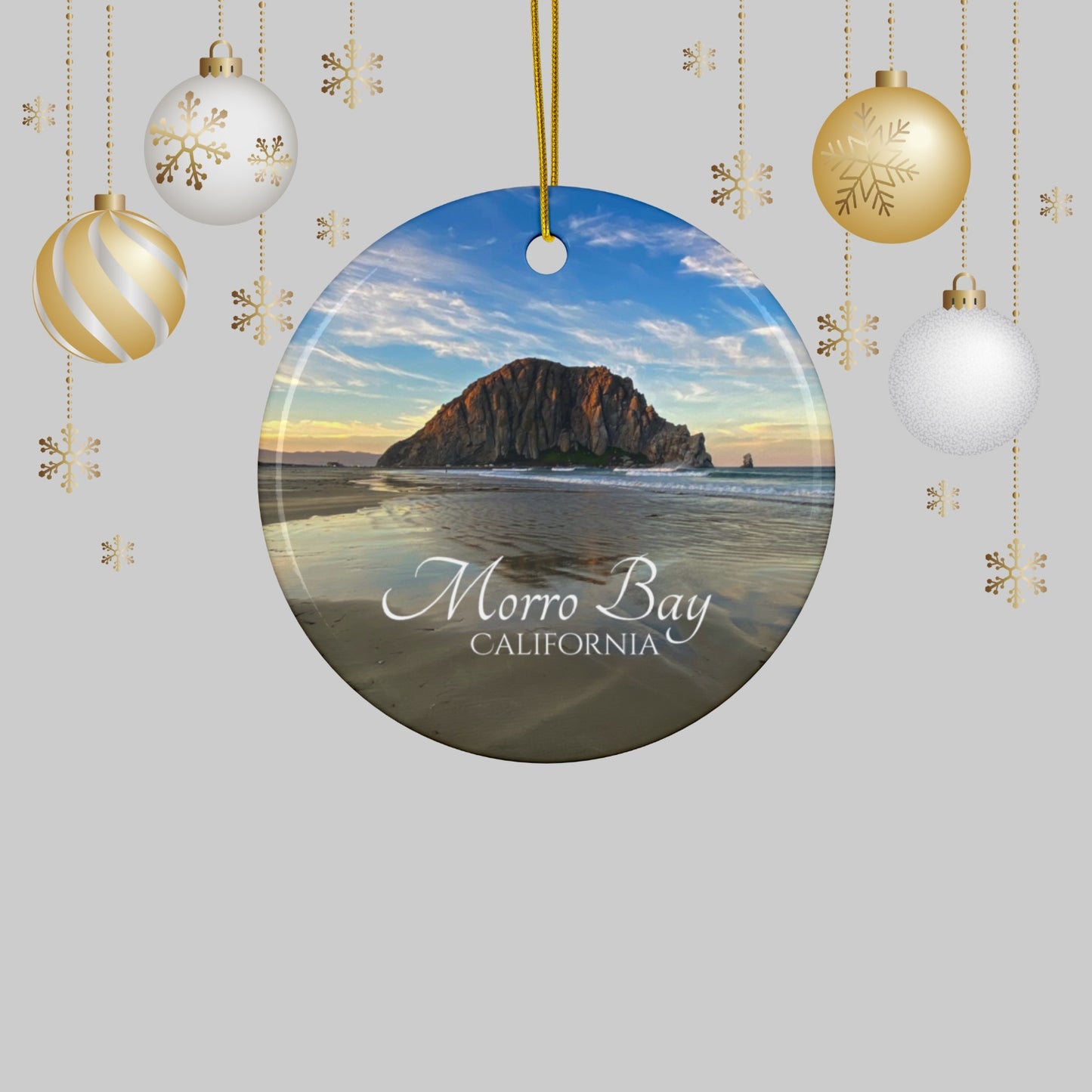 Ceramic holiday ornament featuring a photograph of Morro Rock in Morro Bay, California, with reflections in the sand under a dramatic afternoon sky on a light gray background