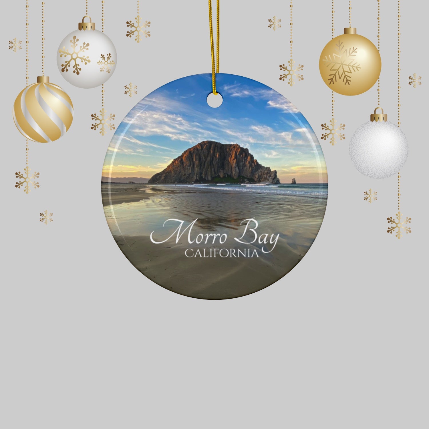 Ceramic holiday ornament featuring a photograph of Morro Rock in Morro Bay, California, with reflections in the sand under a dramatic afternoon sky on a light gray background