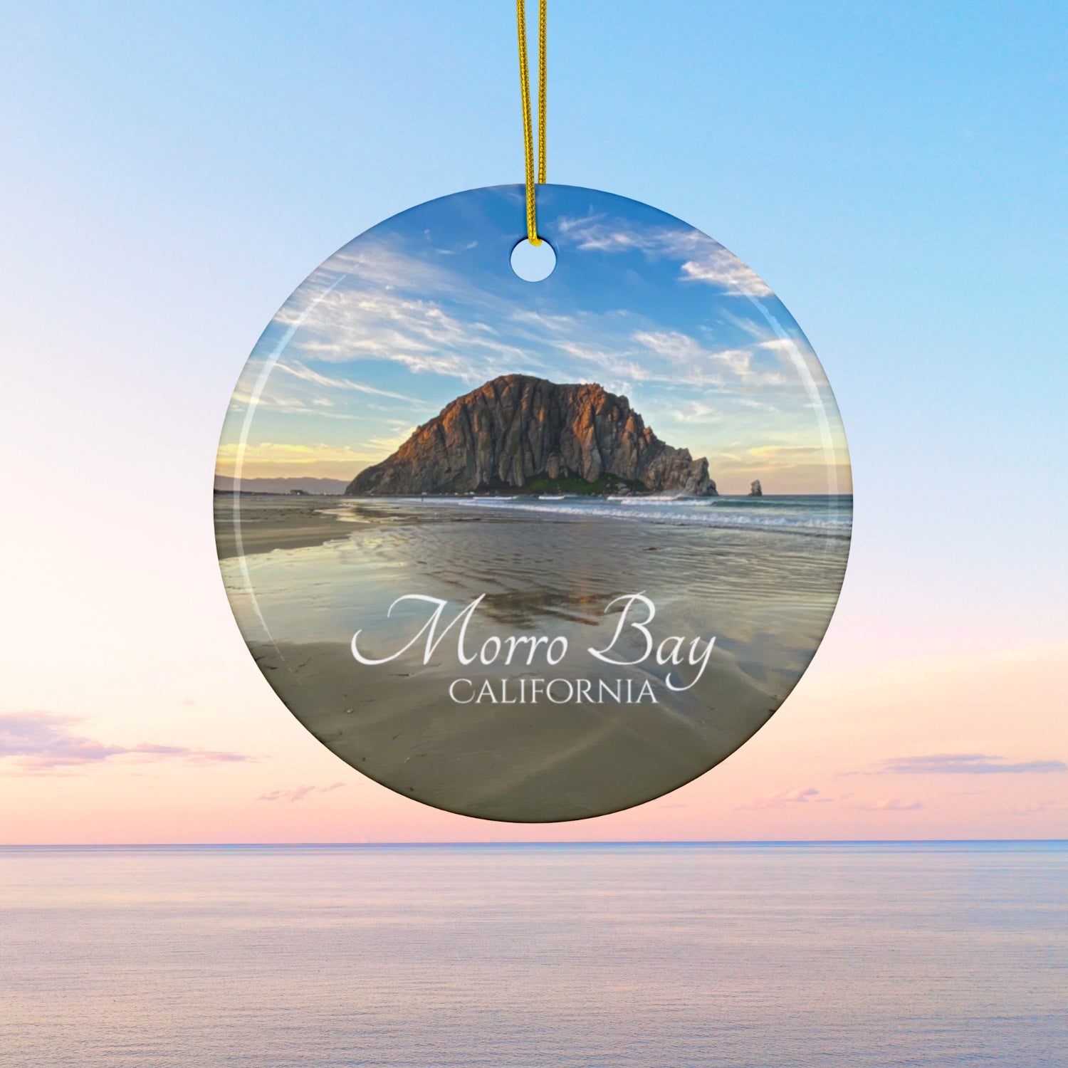 Ceramic holiday ornament featuring a photograph of Morro Rock in Morro Bay, California, with reflections in the sand under a dramatic afternoon sky.