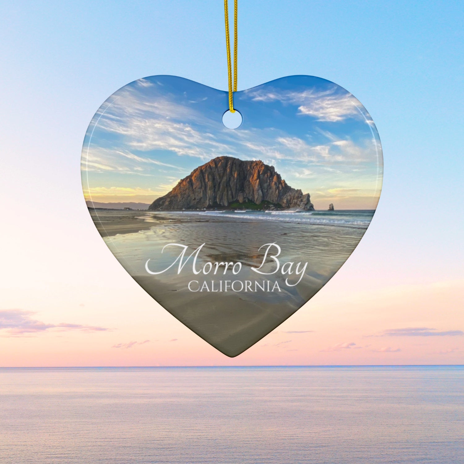 Heart-shaped ceramic holiday ornament featuring a photograph of Morro Rock in Morro Bay, California, with reflections in the sand under a dramatic afternoon sky.