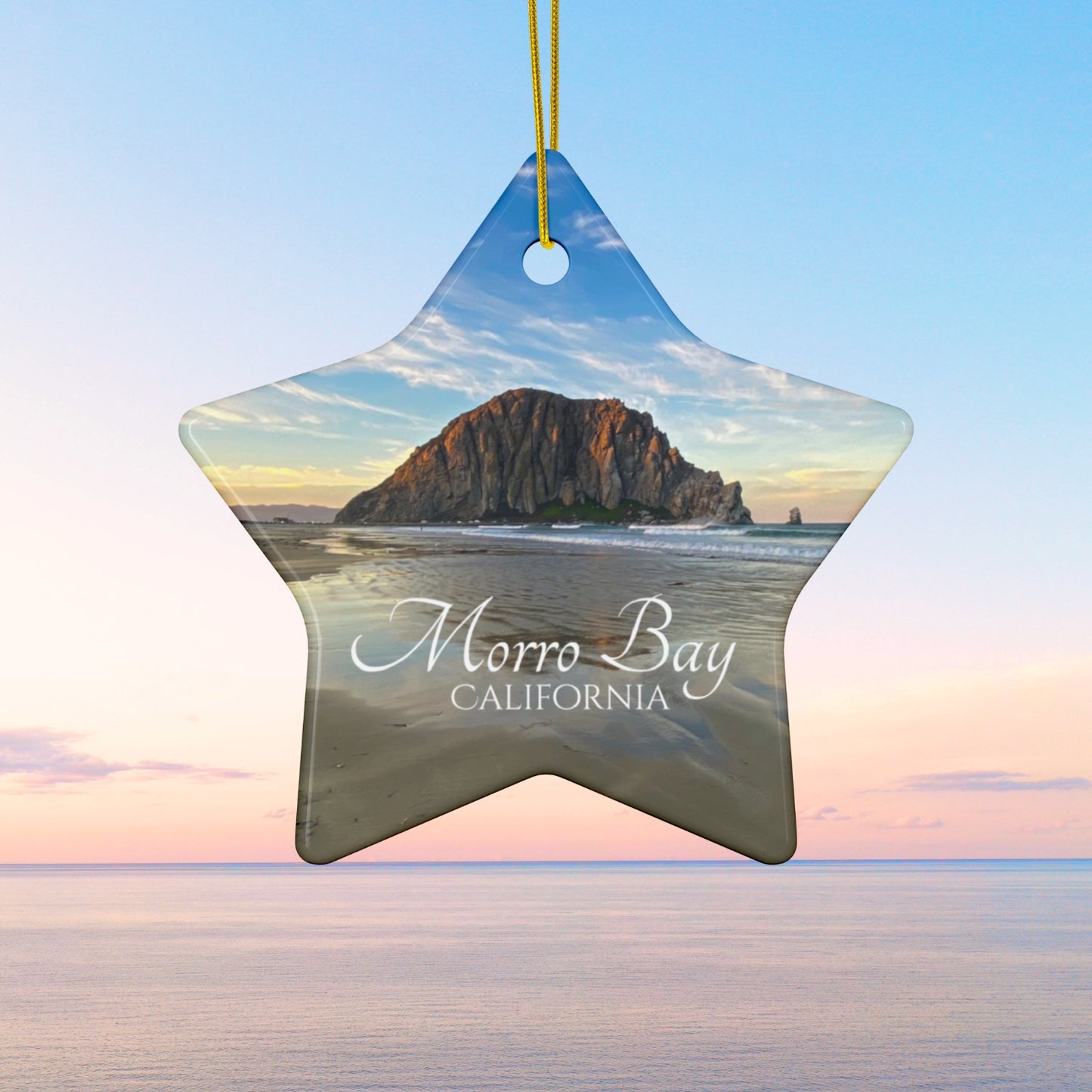 Star-shaped ceramic holiday ornament featuring a photograph of Morro Rock in Morro Bay, California, with reflections in the sand under a dramatic afternoon sky.