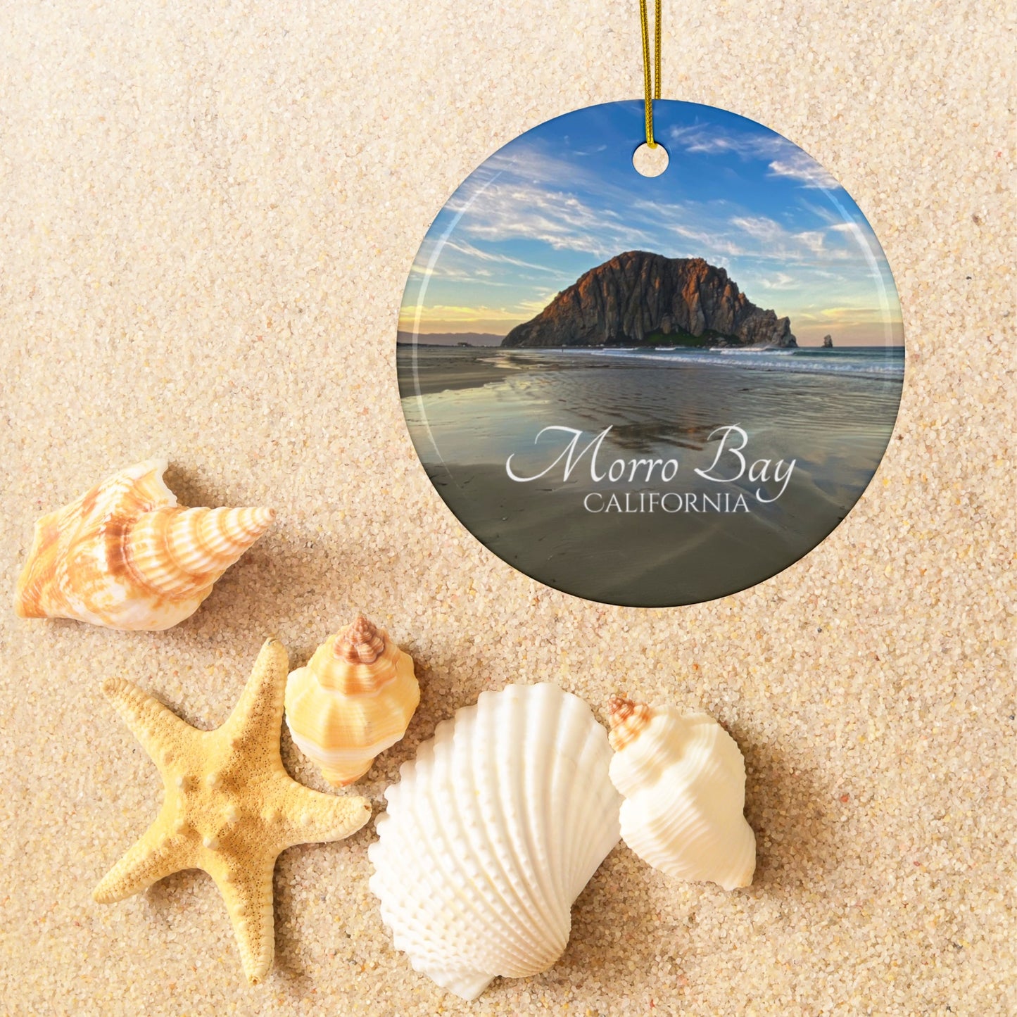 Ceramic holiday ornament featuring a photograph of Morro Rock in Morro Bay, California, with reflections in the sand under a dramatic afternoon sky on a sandy background with shells and a starfish.