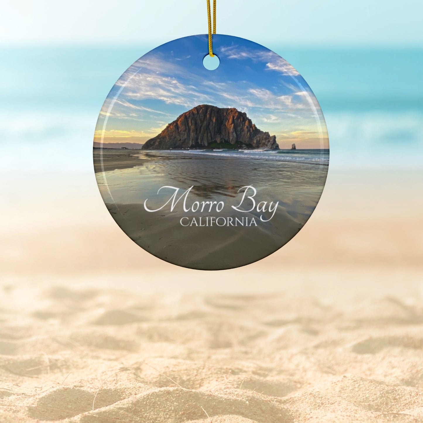Ceramic holiday ornament featuring a photograph of Morro Rock in Morro Bay, California, with reflections in the sand under a dramatic afternoon sky.