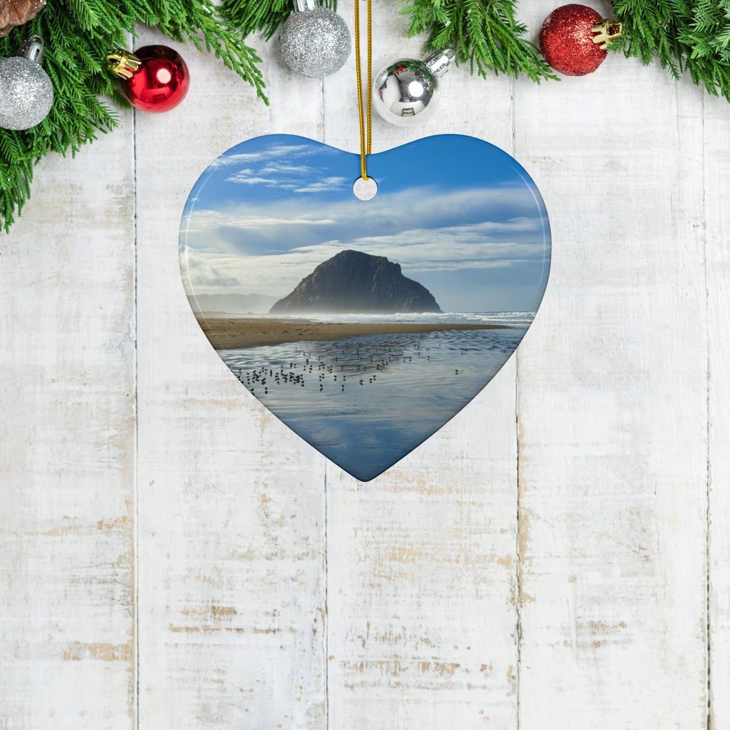 Heart-shaped ceramic holiday ornament featuring a professional photograph of Morro Rock in Morro Bay, California, with the Pacific surf in the foreground, on a wooden background with ornaments.