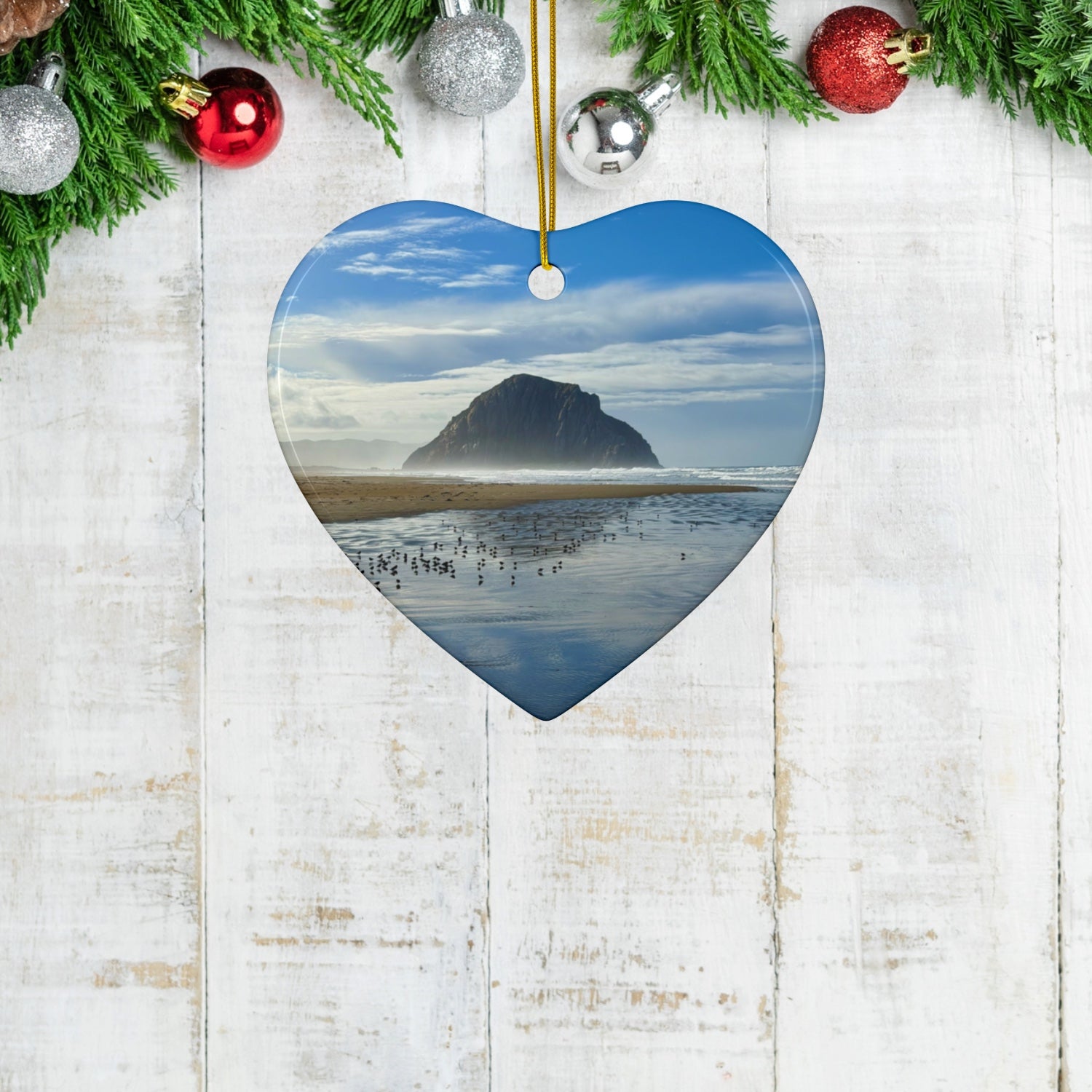 Heart-shaped ceramic holiday ornament featuring a professional photograph of Morro Rock in Morro Bay, California, with the Pacific surf in the foreground, on a wooden background with ornaments.