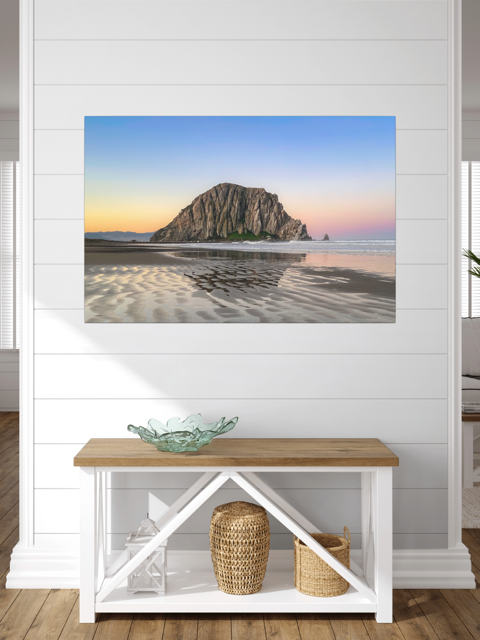 Canvas print featuring a Morro Rock morning reflection in wet beach sand on a wall above a wooden bench with storage baskets.