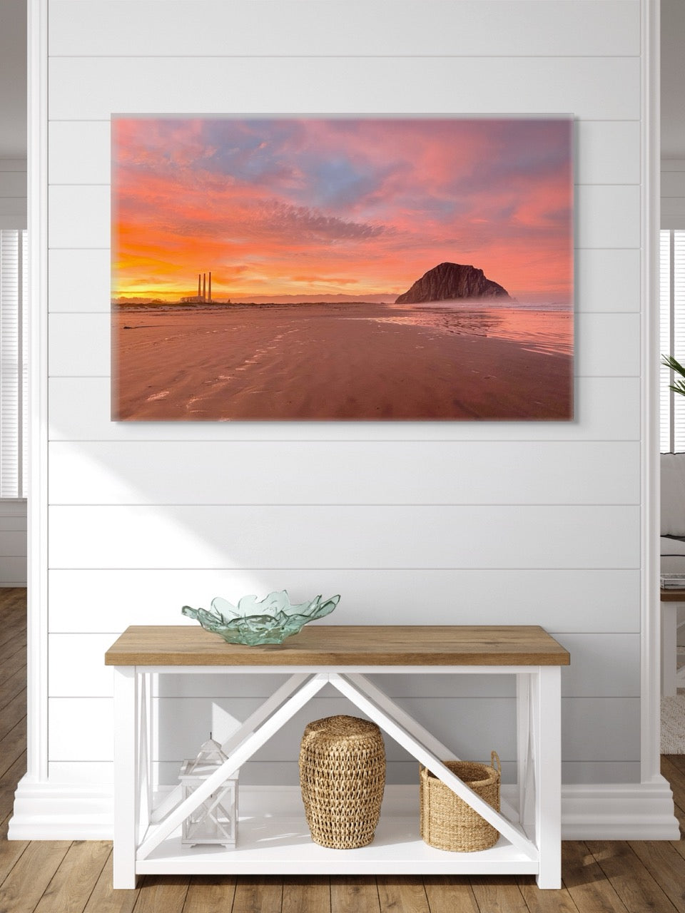 A canvas print of Morro Rock and the stacks at sunrise on a wall above a wooden bench with a glass bowl and woven baskets.