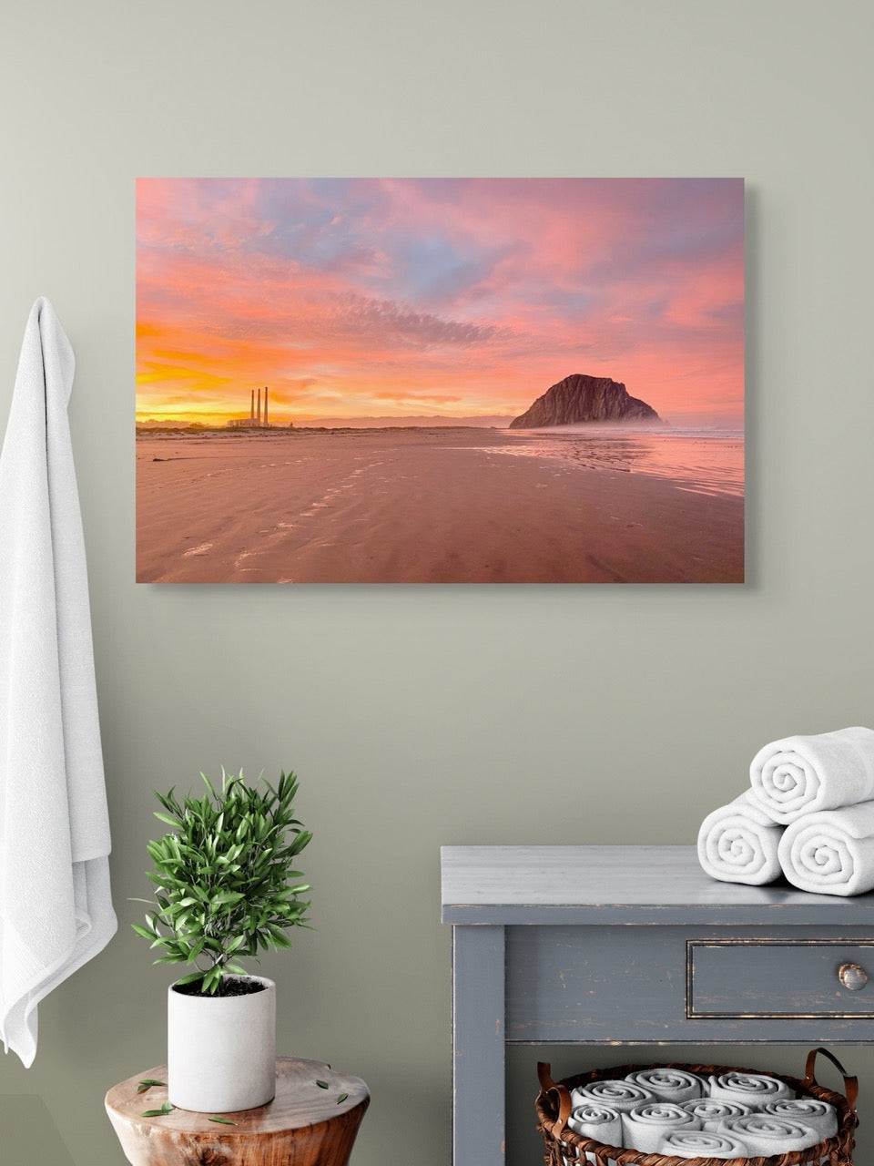 A canvas print of Morro Rock and the stacks at sunrise on a wall above a dresser with towels and a plant.