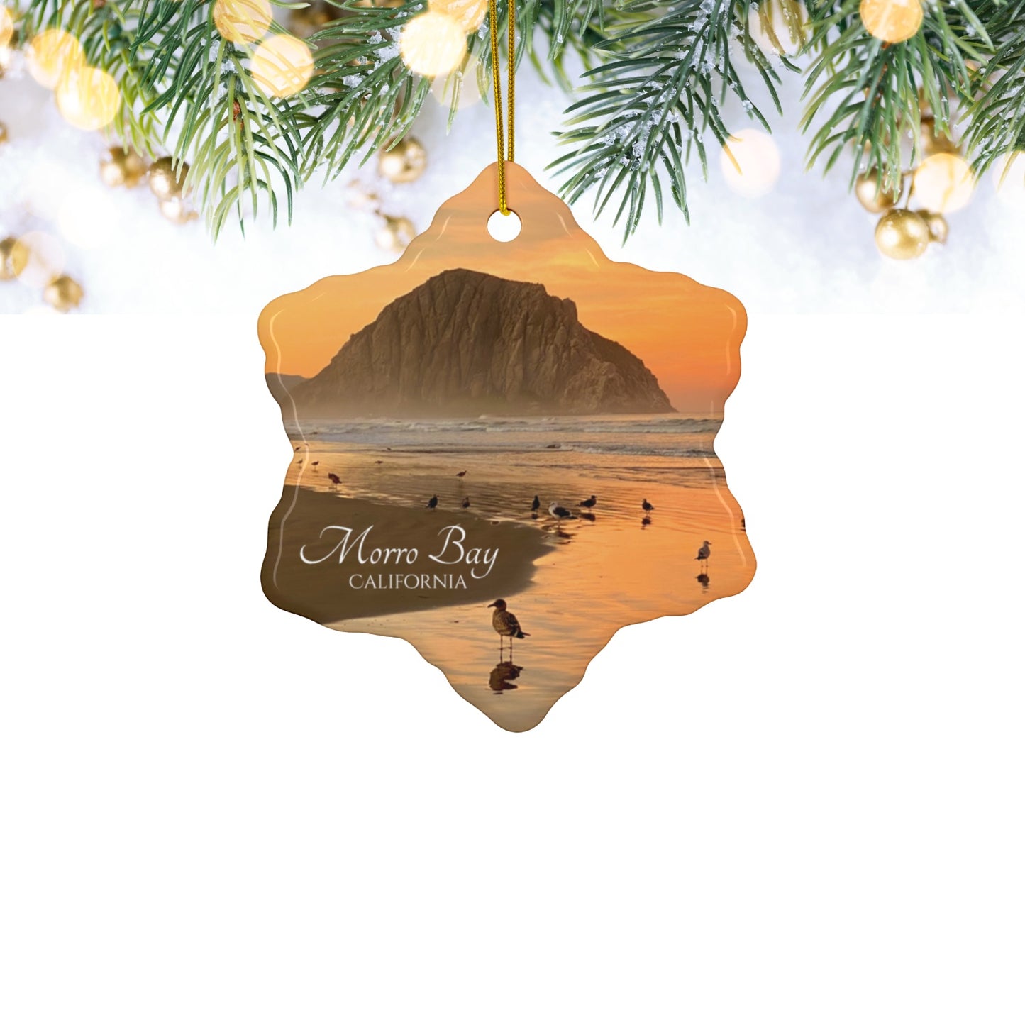 Ceramic Christmas ornament featuring a photograph of a golden sunrise over Morro Rock with shorebirds reflected in the wet sand of the shoreline, hanging against a white background.
