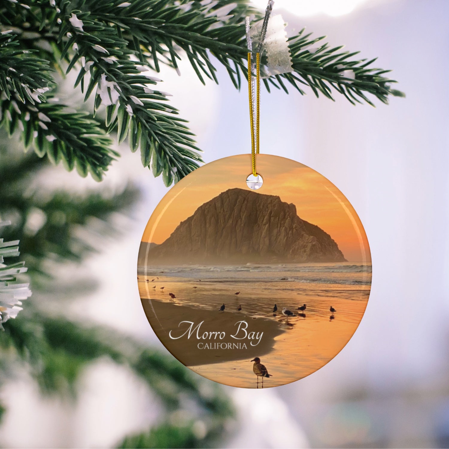 Ceramic Christmas ornament featuring a photograph of a golden sunrise over Morro Rock with shorebirds reflected in the wet sand of the shoreline hanging on a Christmas tree branch.