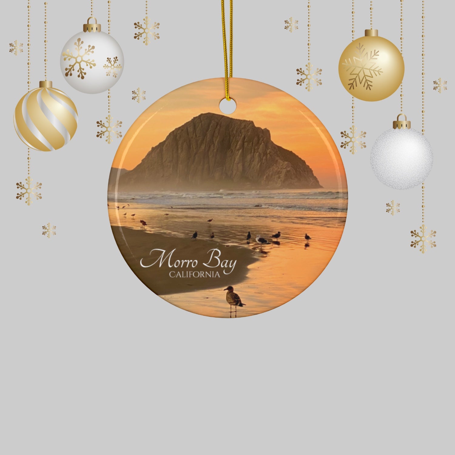 Ceramic Christmas ornament featuring a photograph of a golden sunrise over Morro Rock with shorebirds reflected in the wet sand of the shoreline on a gray background with Christmas ornaments.