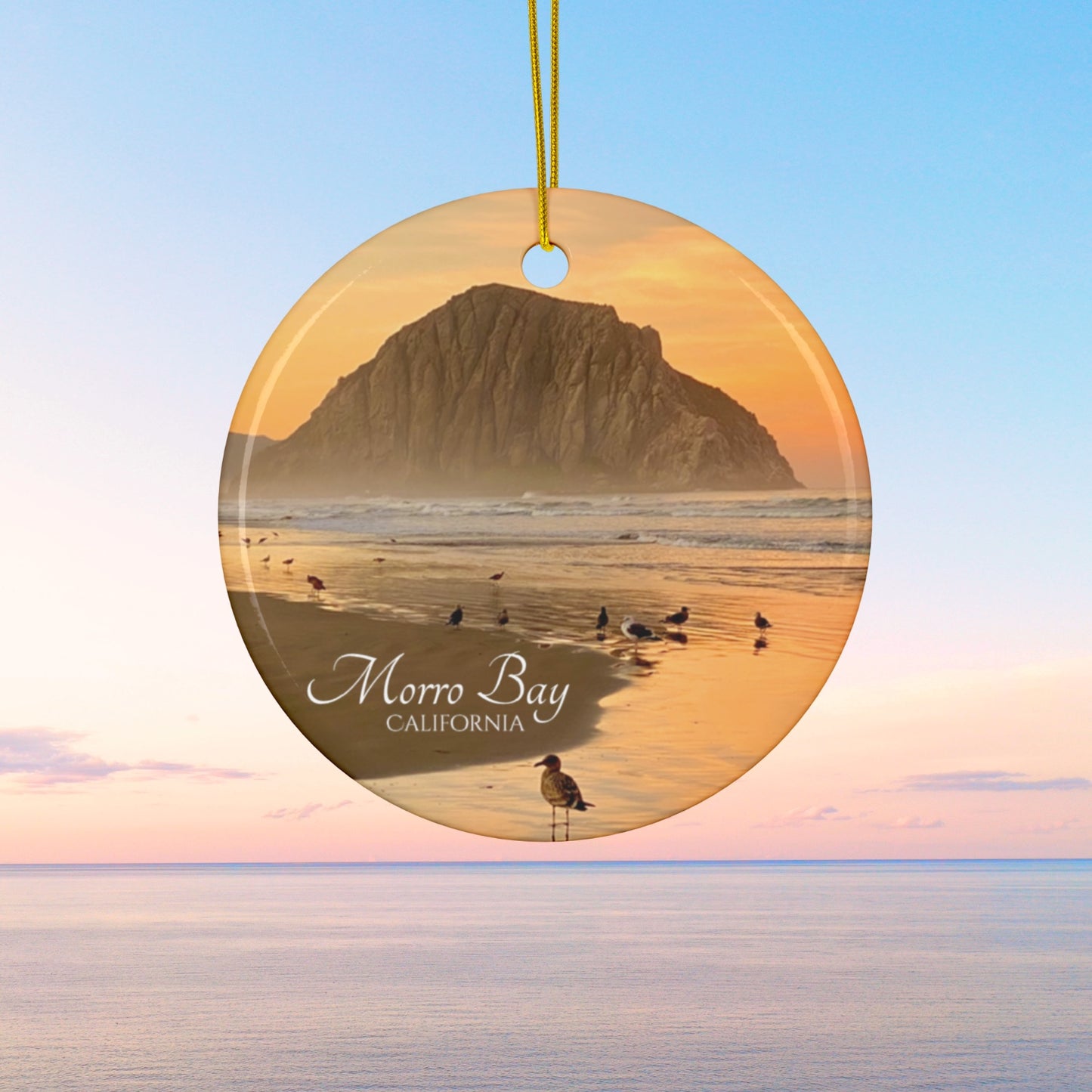 Ceramic Christmas ornament featuring a photograph of a golden sunrise over Morro Rock with shorebirds reflected in the wet sand of the shoreline.