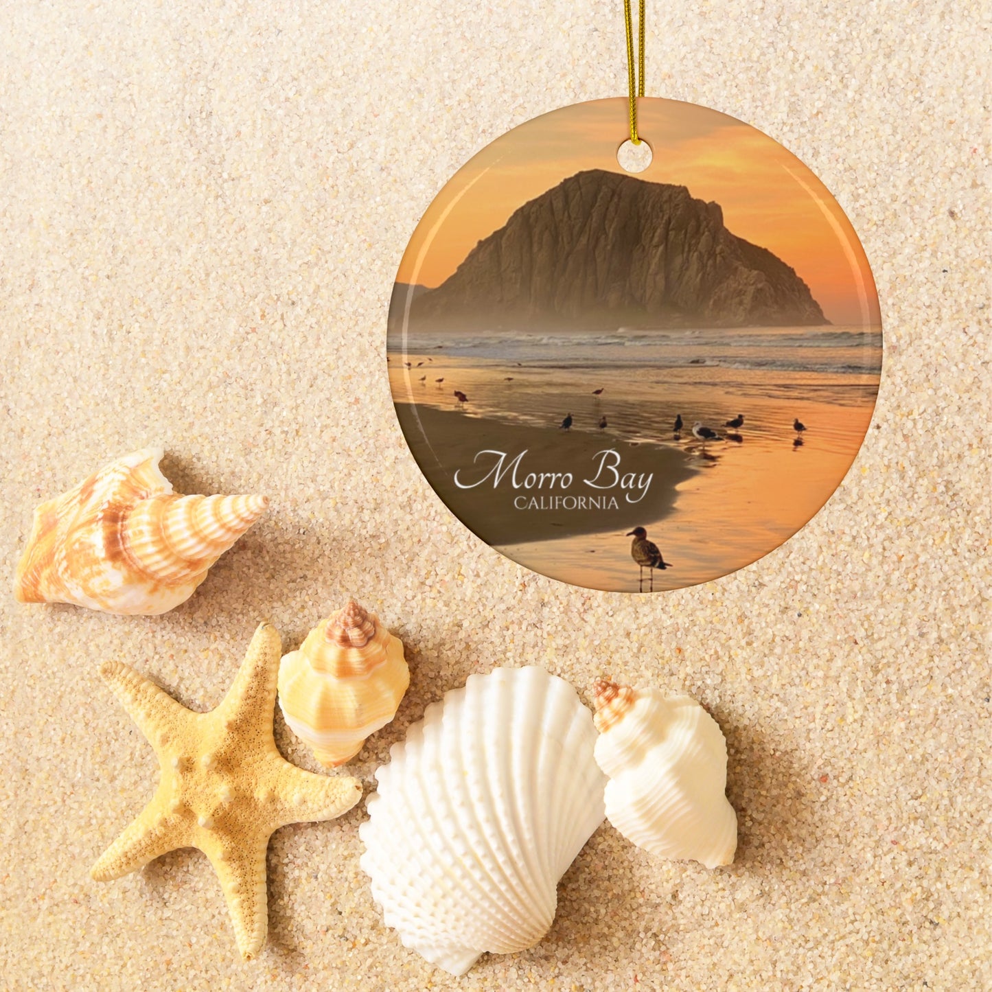 Ceramic Christmas ornament featuring a photograph of a golden sunrise over Morro Rock with shorebirds reflected in the wet sand of the shoreline on a sandy background with shells and a starfish.