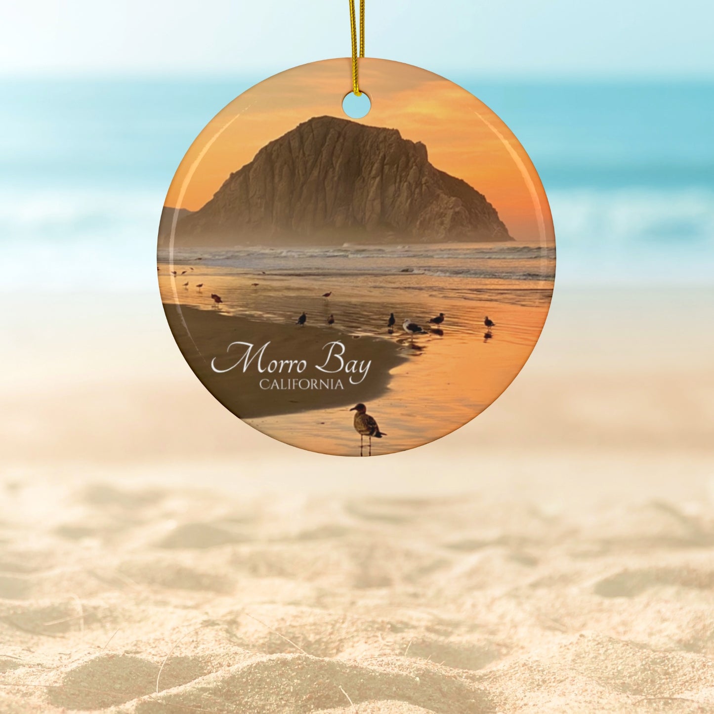 Ceramic Christmas ornament featuring a photograph of a golden sunrise over Morro Rock with shorebirds reflected in the wet sand of the shoreline.