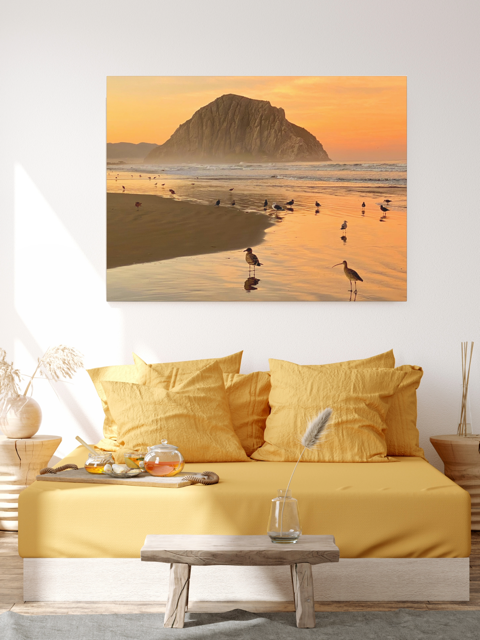 A canvas print featuring an orange colored sunrise sky over Morro Rock with birds on the shoreline in the foreground above a yellow bed in a bedroom