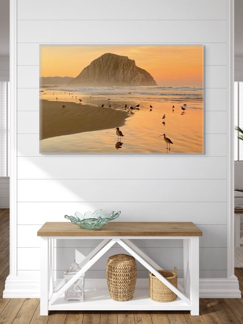 A canvas print featuring an orange colored sunrise sky over Morro Rock with birds on the shoreline in the foreground on a wall above a wooden bench with a glass bowl and baskets.