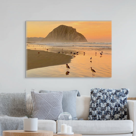 Large canvas print featuring an orange colored sunrise sky over Morro Rock with birds on the shoreline in the foreground on a wall above a sofa.