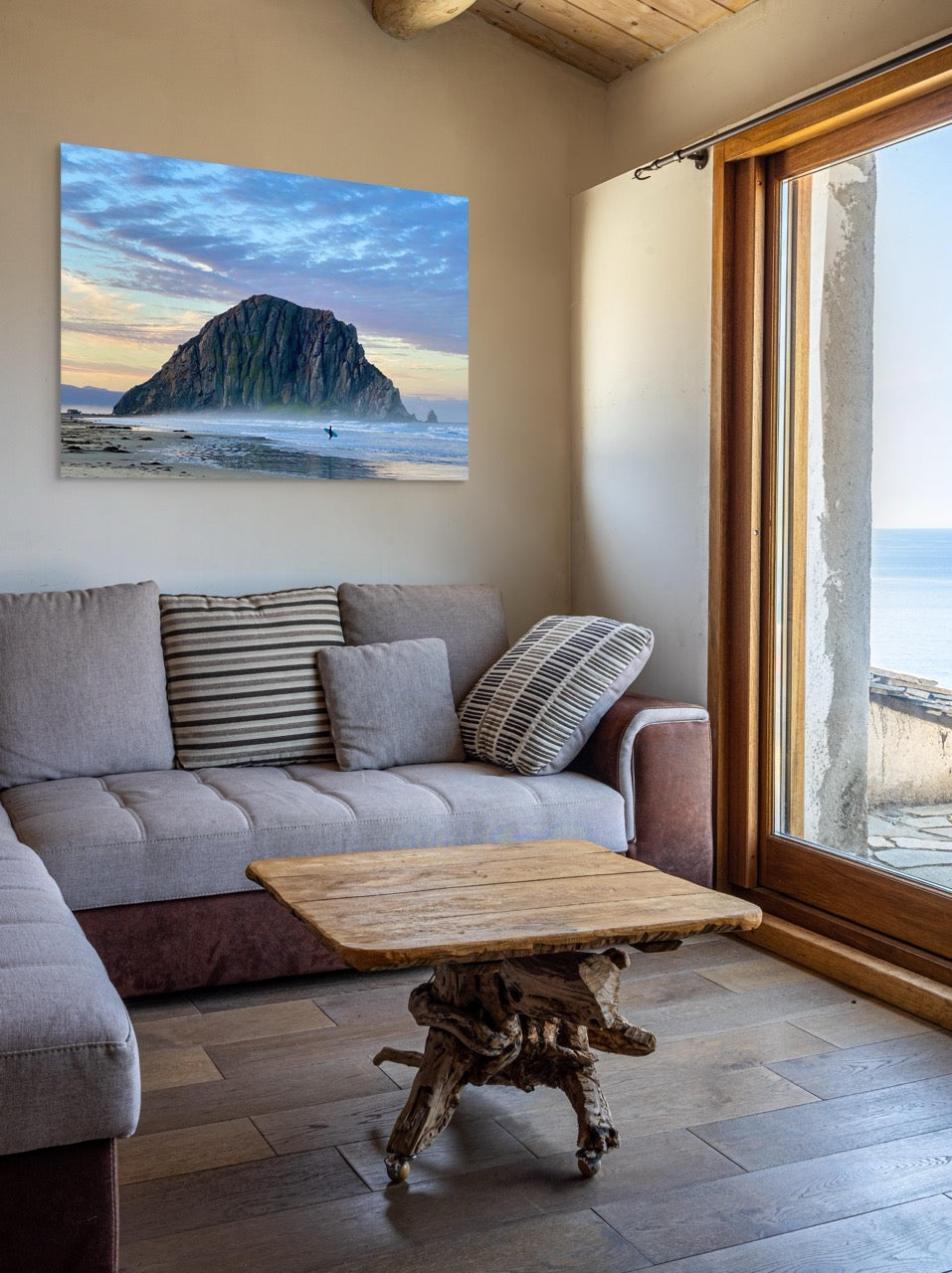 Canvas print of a sunrise at Morro Rock in Morro Bay, California with a surfer on the beach in a living room with a gray sofa, wooden coffee table, and a large window showing a coastal view.