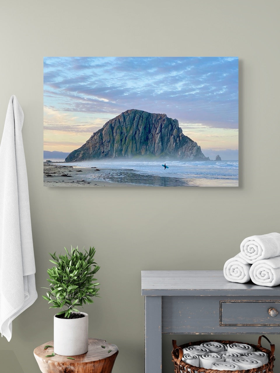 Canvas print of a sunrise at Morro Rock in Morro Bay, California with a surfer on the beach on a light green wall above a gray cabinet.