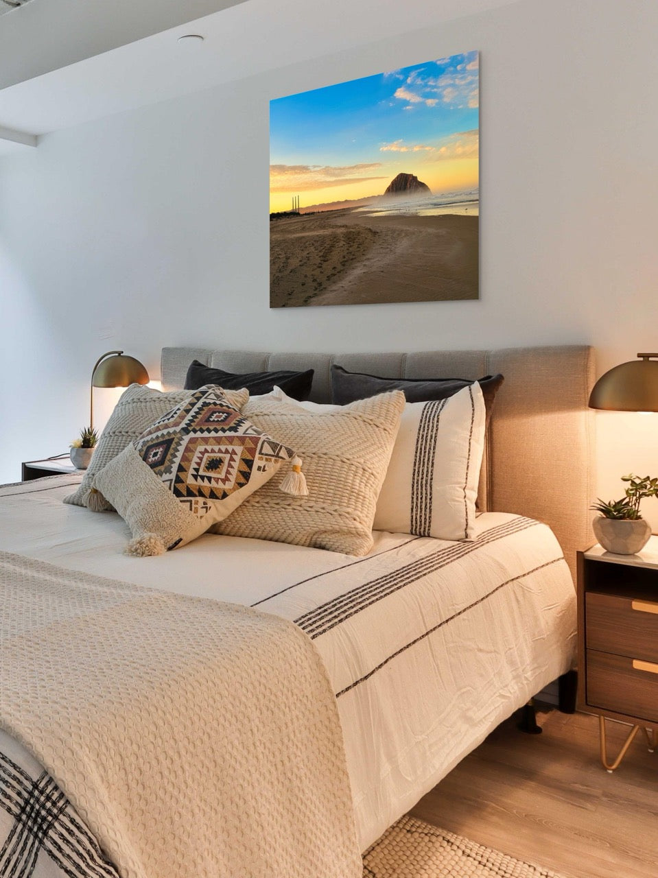 Bedroom with a large bed and decorative pillows, featuring a canvas print of Morro Rock and the stack at sunrise on the wall.
