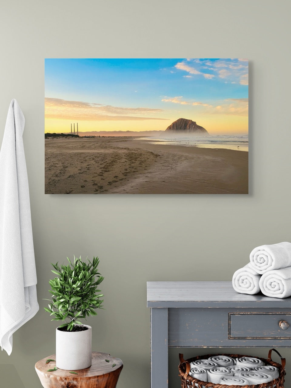 Canvas print of Morro Rock and the stack at sunrise on a wall above a table with a plant and folded towels.