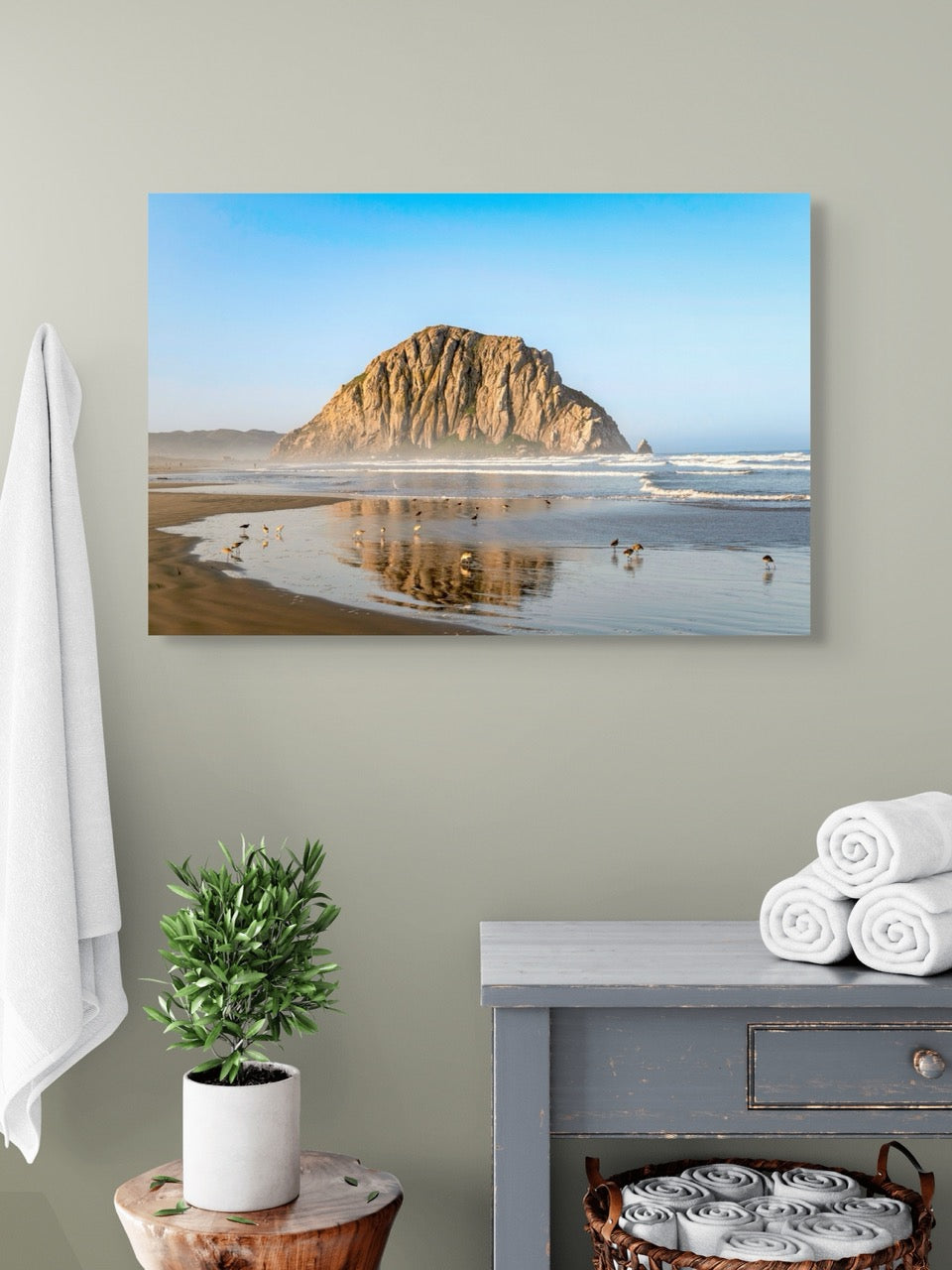 Canvas print of Morro Rock reflecting in the wet sand with birds on the shoreline on a wall above a table with towels and a plant.