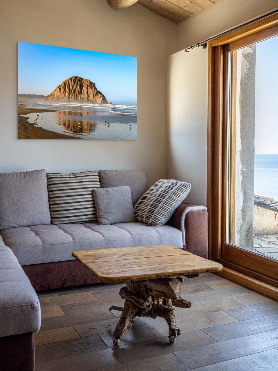Living room with a gray sofa, wooden coffee table, and a large canvas print of Morro Rock reflecting in the wet sand with birds on the shoreline.