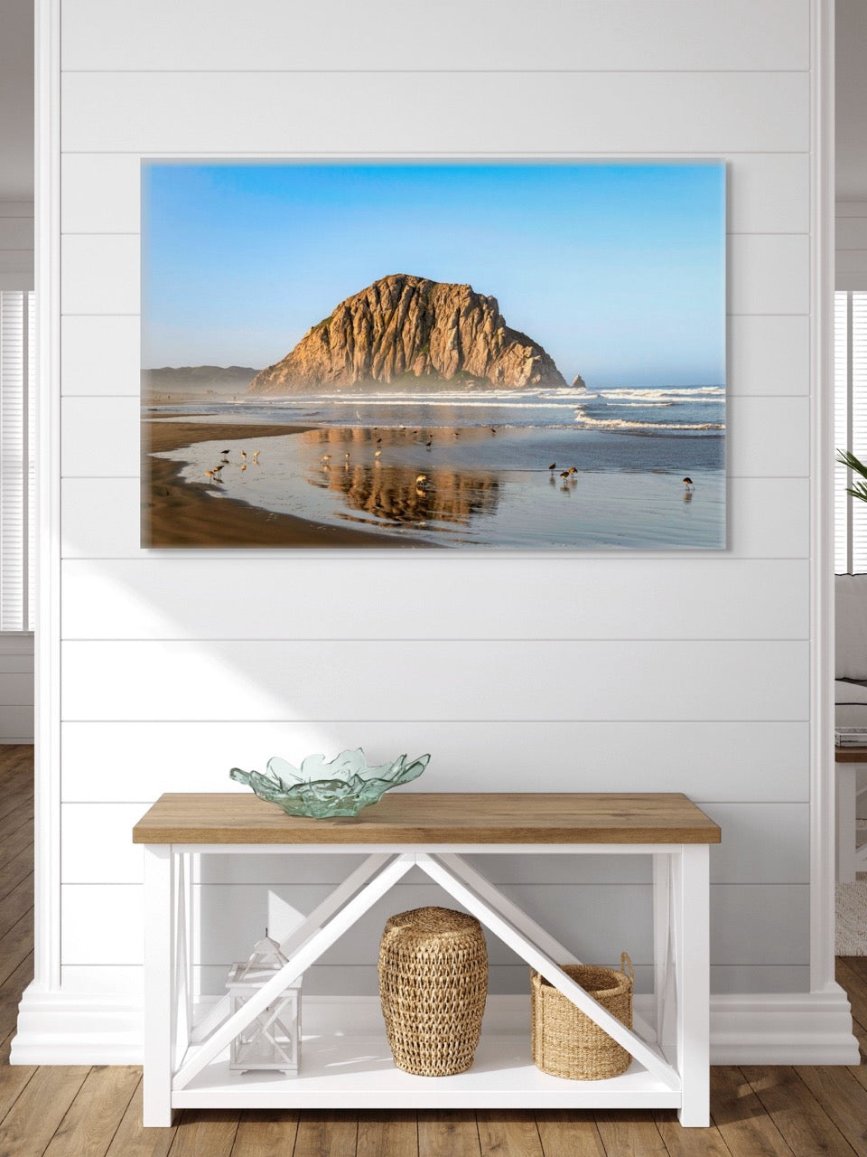A canvas print of Morro Rock reflecting in the wet sand with birds on the shoreline on a wall above a wooden bench with storage.