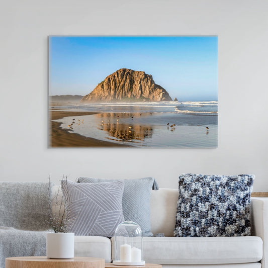 A canvas print of Morro Rock reflecting in the wet sand with birds on the shoreline on a wall above a sofa with decorative pillows.