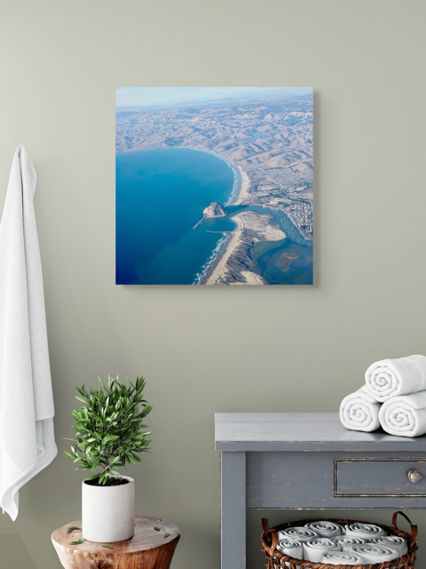 Aerial photography of Morro Rock and California coastline canvas print on a wall in a room with a plant and folded towels.